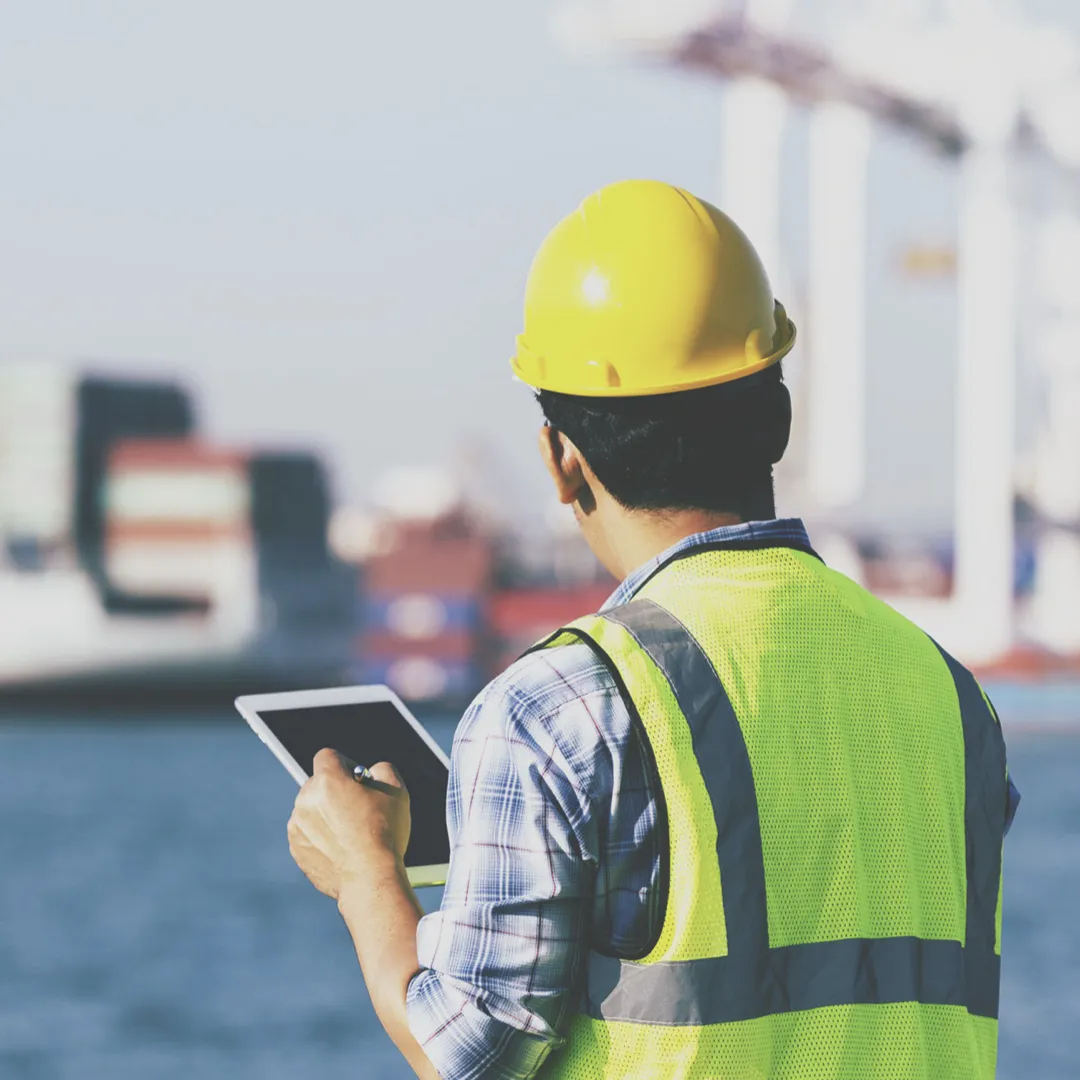 Inspecteur met helm en tablet controleert een schip in de haven, gericht op veiligheid en chemisch beheer aan boord.